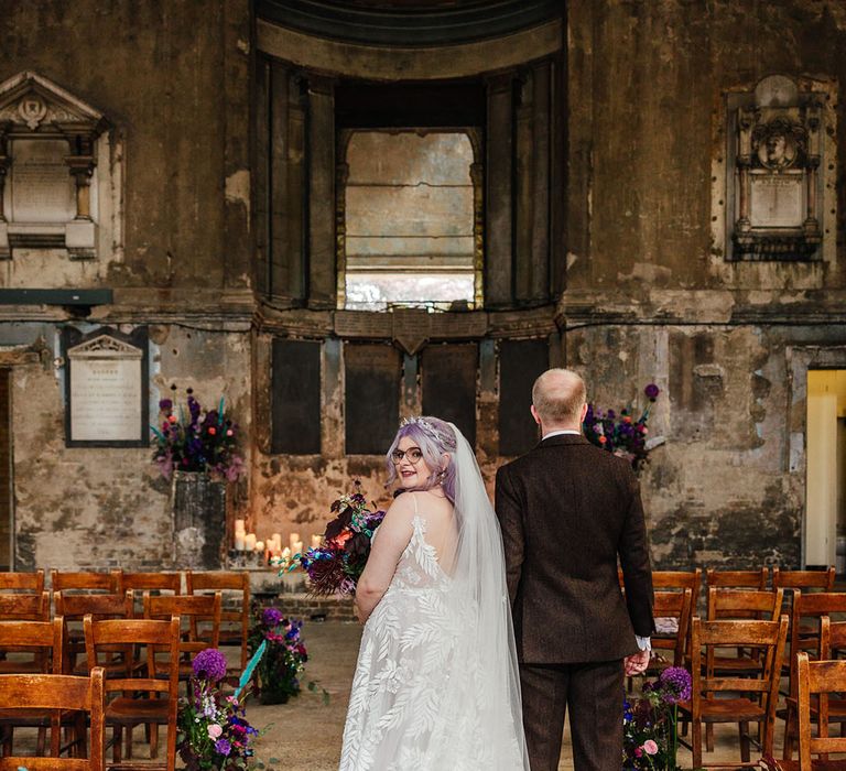 Groom in mocha brown textured suit standing with bride in leaf appliqué sheer Evie Young bridal gown and cathedral length veil at Asylum Chapel London wedding venue 