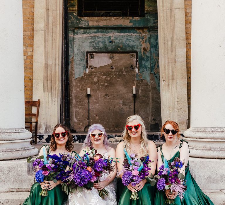 Bride in leaf appliqué sheer Evie Young bridal gown and cathedral length veil and white heart shaped wedding sunglasses and holding bright purple bridal bouquet with purple hydrangeas, pink garden roses, purple orchids, lilac carnations, dried flowers and peacock blue feathers sitting with bridesmaids in satin forest green bridesmaid dresses and heart shaped sunglasses 