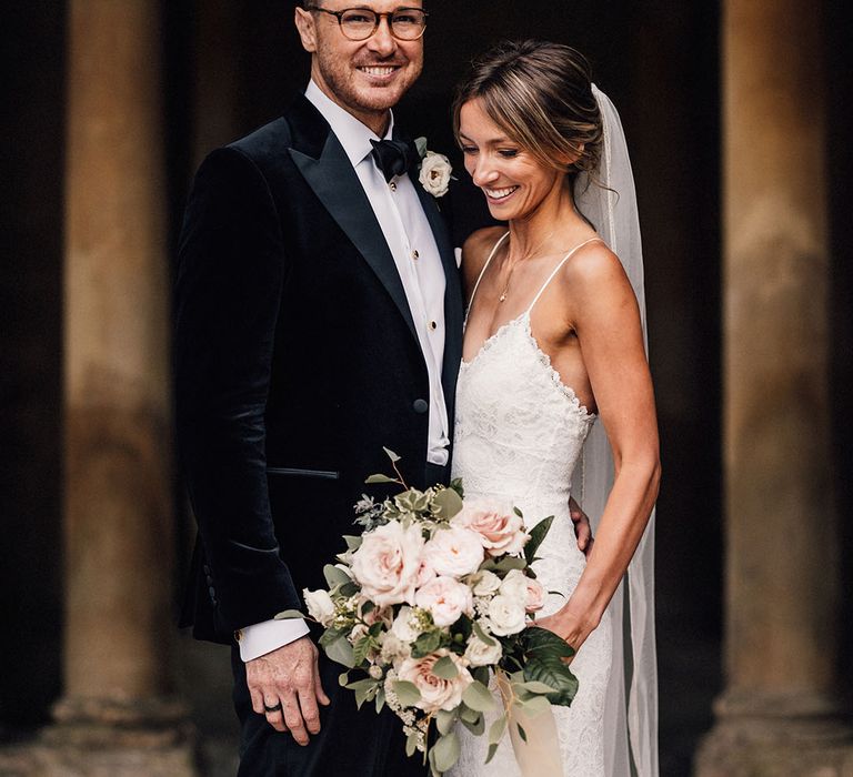 The groom wears glasses in a black velvet tuxedo with the bride in a fitted lace wedding dress holding a pink rose bouquet 