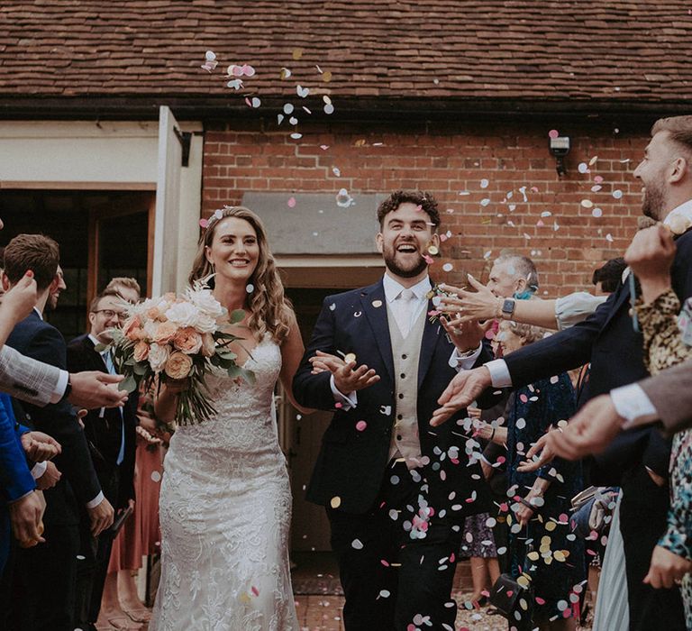 The bride has a confetti moment with the bride in a lace fitted wedding dress with the groom in a navy suit and light coloured waistcoat 