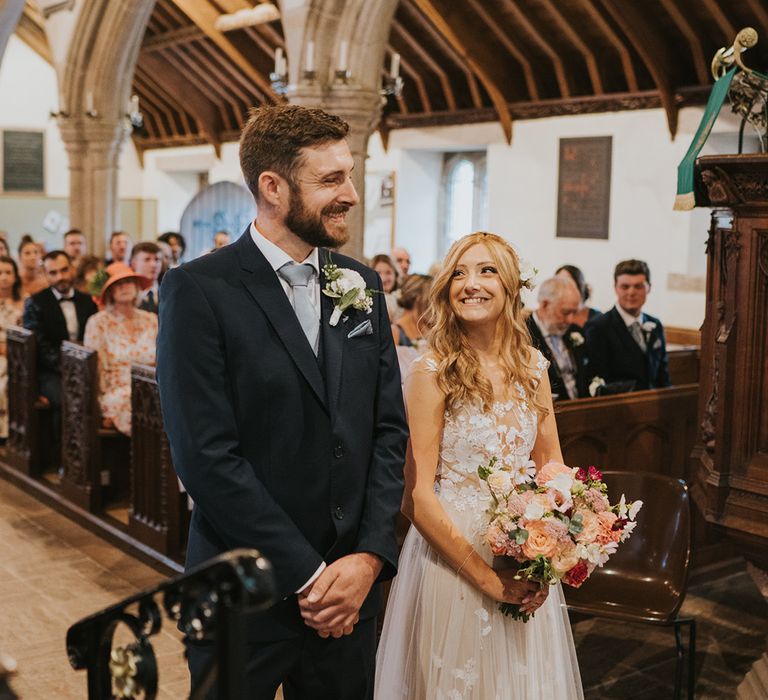 Smiling groom in navy suit with light blue tie and white flower buttonhole with the bride in lace wedding dress and flower hair accessory standing at the altar for the wedding ceremony 