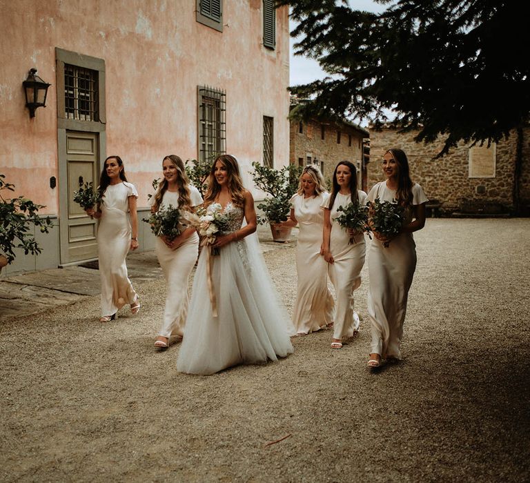 Bride in embroidered tulle wedding dress walking with bridesmaids in off-white satin bridesmaids dresses at italian wedding villa holding eucalyptus bouquets