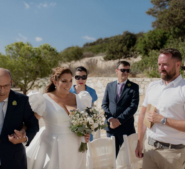 Bride in puff-sleeve satin white wedding dress walks into wedding ceremony with father of the bride