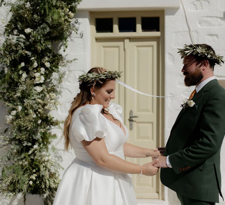 Bride and groom wearing green floral crowns and holding hand at intimate wedding ceremony with green and white flower column decor