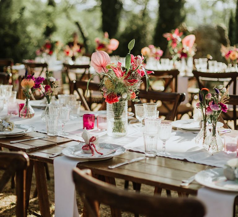 Rustic wooden tables with white and bright pink tablescape featuring vases of peonies, anthuriums, and bright pink flowers at woodland wedding in Italy