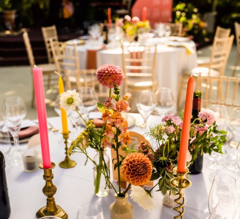 Orange and pink dahlias in bud vases with colourful orange, pink, and yellow candles in gold sculptural candle holders 