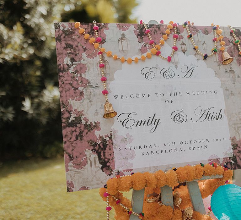 Colourful Indian wedding welcome sign with black cursive lettering, orange garlands, blue lanterns and golden bells 