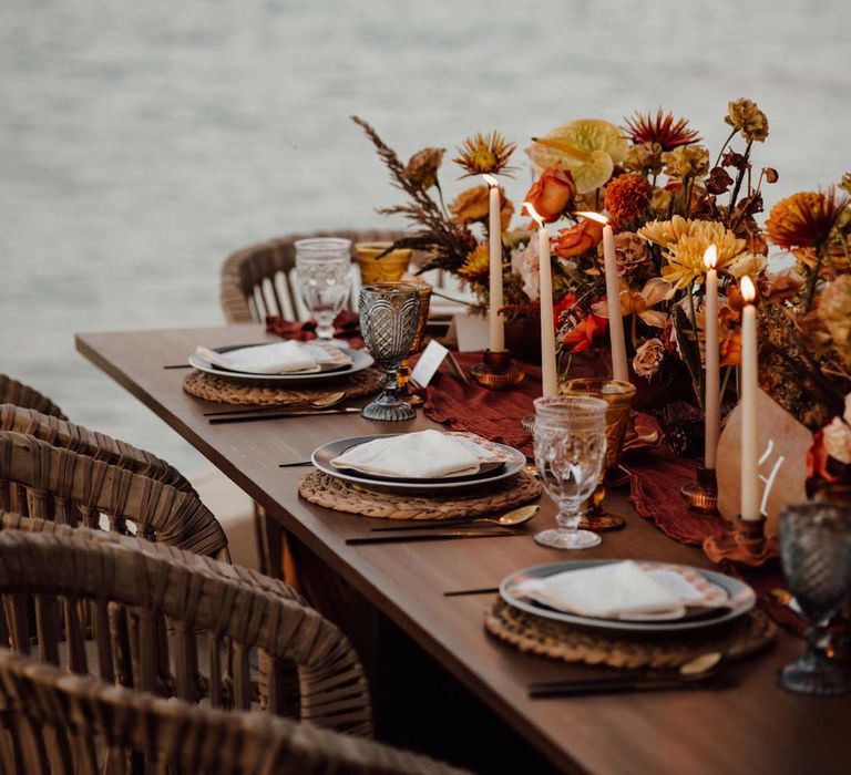 Rustic wedding tablescape by the Mexican sea with wicker chairs, burnt orange dried roses, dried flower and foliage floral centrepieces, wooden slate wedding table numbers, light orange tapered candles and burnt orange table runners