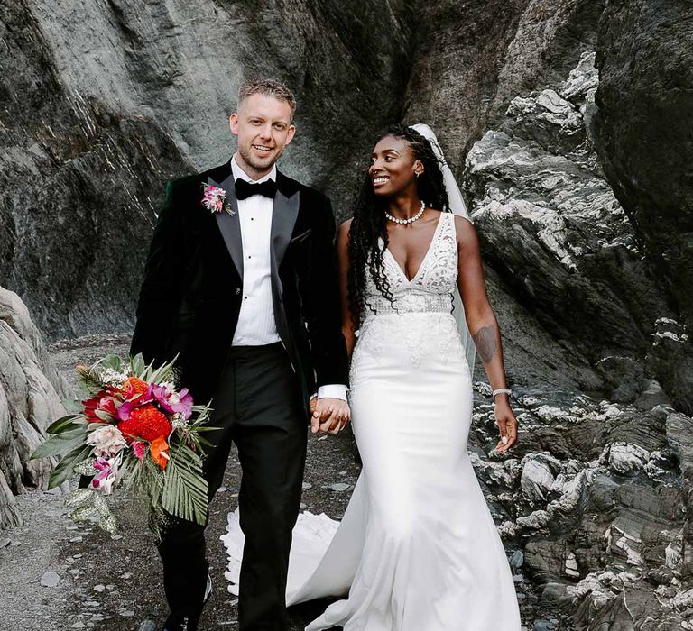 Groom in bottle green velvet grooms blazer, colourful tropical boutonniere and black suit trousers holding colourful tropical African bridal bouquet and holding hands with bride in v neck sleeveless lace Pronovias wedding dress, cathedral length veil and silver sparkly bridal jewellery 