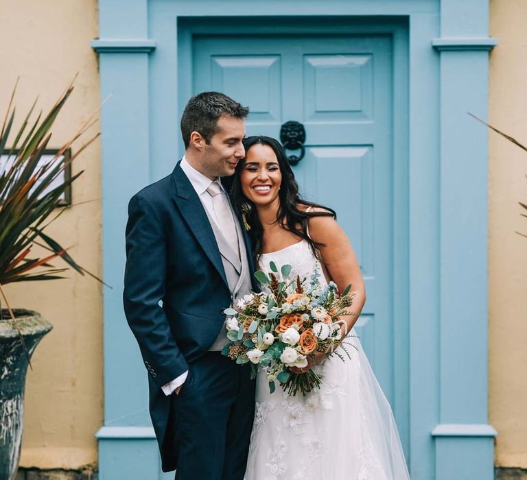 Bride in a-line tulle wedding dress with beaded lace applique with petals and a square neckline with side tulle cutouts holding pampas grass, eucalyptus, blush garden roses, dried flowers and foliage bridal bouquet embracing groom in navy grooms morning suit with grey waistcoat, off-white tie and mixed wildflower boutonniere standing in front of baby blue door at South Farm wedding venue