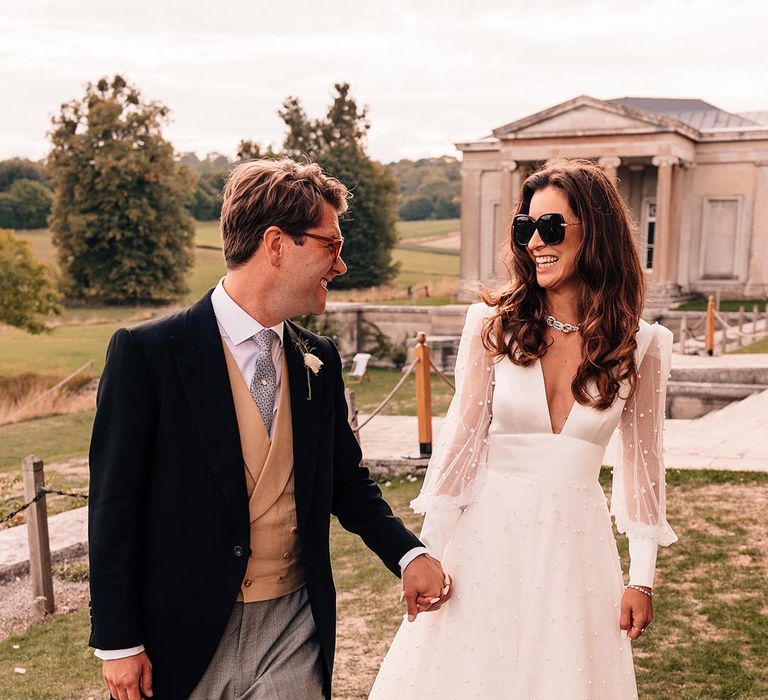 The groom in a traditional morning suit with the bride in a pearl wedding dress with long sleeves wearing sunglasses 