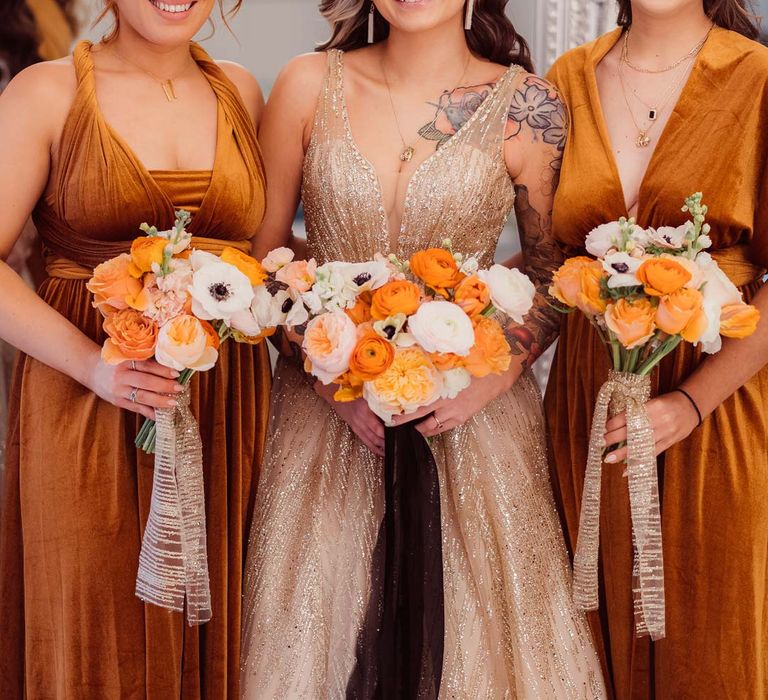 Bride in sleeveless deep v-neck sparkly gold wedding dress with sheer and gold polka dot wedding veil standing with bridesmaids in velvet burnt orange bridesmaid dresses all holding orange garden rose, white poppy, light pink carnation and foliage bridal bouquets wrapped in sparkly gold ribbon