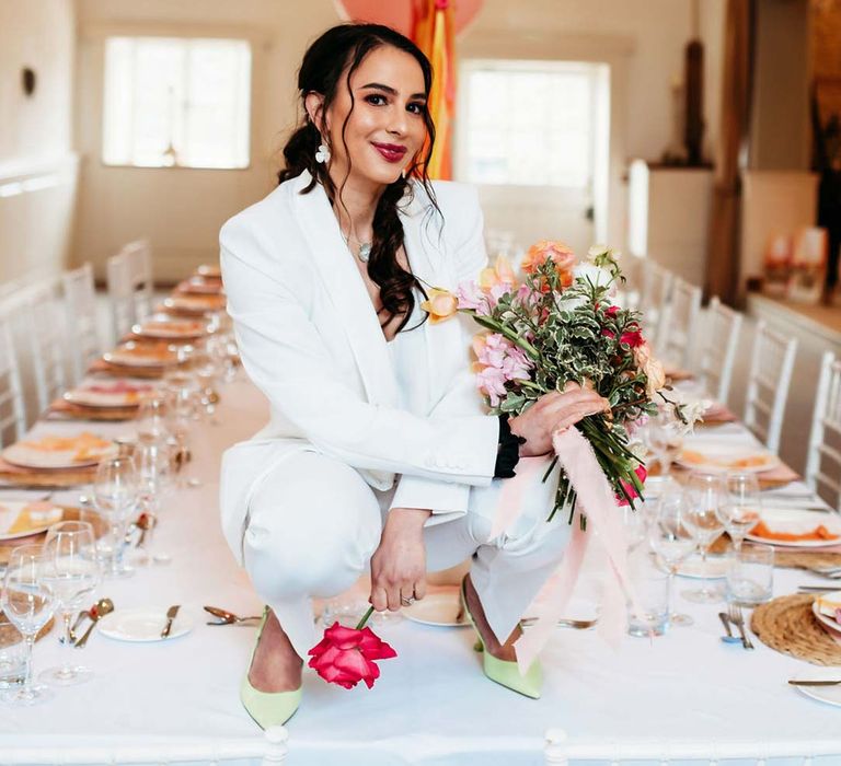 Bride white bridal suit with silver bridal jewellery with closed toe bright green wedding heels squatting on wedding tablescape with white wedding tablecloth, burlap place mats, tapered candles, pink and orange wedding stationery, and floral wedding table centrepieces with pink and orange garden roses, orange floribunda, yellow ranunculus flowers, foliage and Iceland poppies 