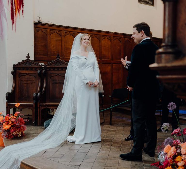 Bride in asymmetrical off shoulder long sleeve satin wedding dress with cathedral length personalised veil standing at the alter of Fabrica Brighton wedding venue with groom 