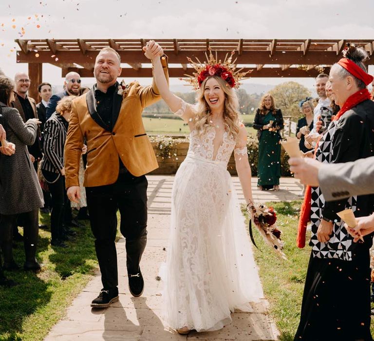 Groom in crushed velvet burnt orange grooms blazer and dried flower boutonniere holding hands with bride in off shoulder lace wedding dress with corset style top and red rose and gold halo bridal crown holding red rose, pampas grass and dried flower bridal bouquet doing colourful confetti exit at Southlands Barn wedding venue