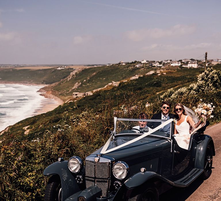 bride and groom riding in a vintage wedding car with views of Sennen Cove behind them