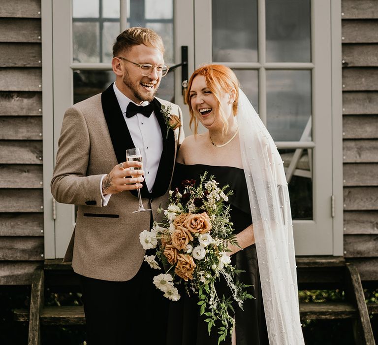 Bride carrying cascading wedding bouquet with gold necklace in black wedding dress laughing with the groom drinking champagne 