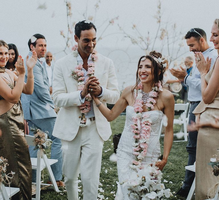 Bride and groom walk through confetti exit holding a white orchid wedding bouquet and wearing traditional pink flower varmalas