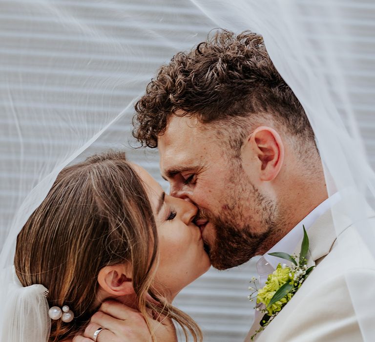 The bride sits wearing pearl hair clips kissing the groom underneath the wedding veil 