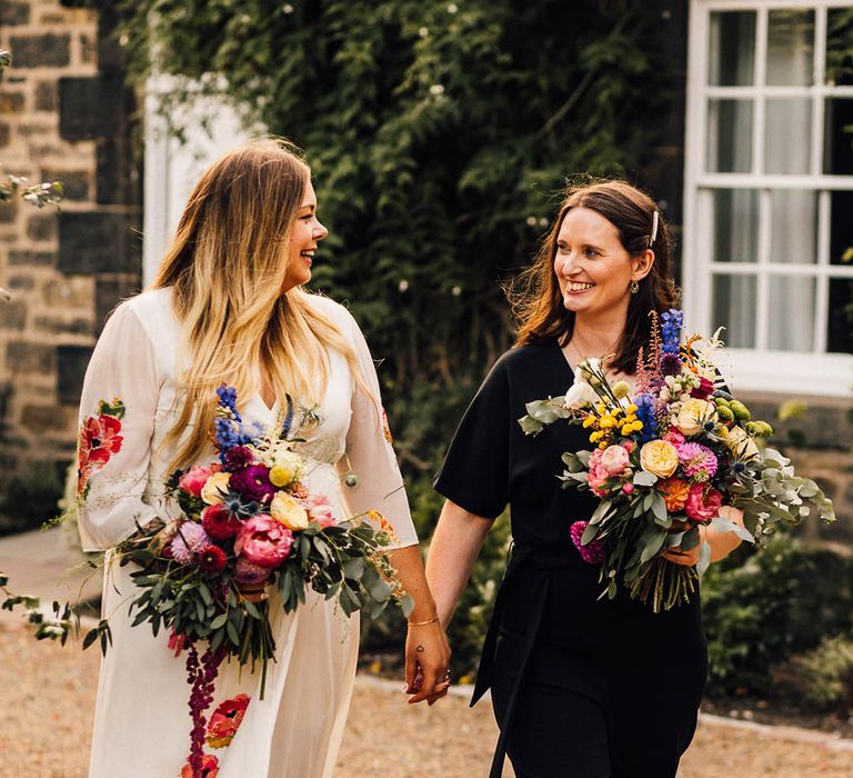 Two brides at same sex wedding with bride wearing colourful embroidered wedding dress and bride in bridal black jumpsuit holding colourful bouquets