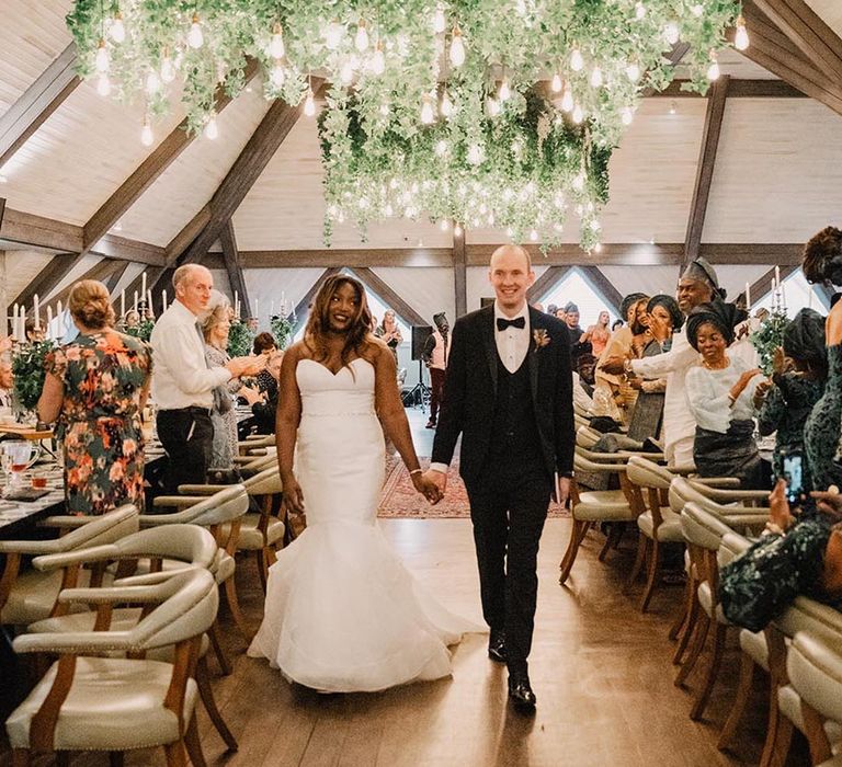 Bride in strapless mermaid wedding dress walking with groom in black tuxedo walking together at wedding reception 