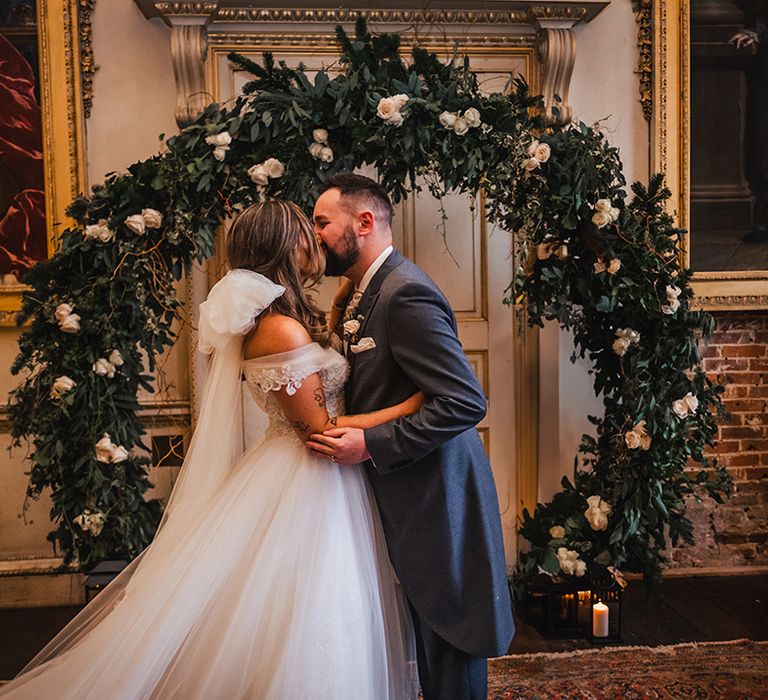 Bride and groom share their first kiss as a married couple in front of white flower arch decoration 