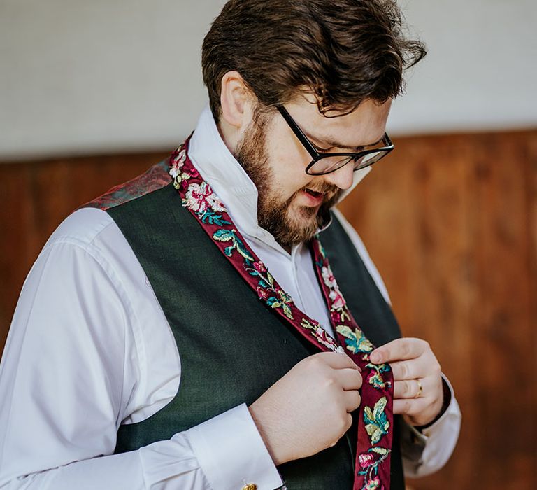 Groom in a white and black tuxedo putting on red floral embroidered tie 