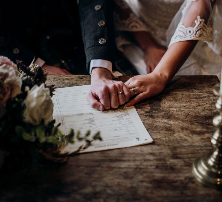 Scottish boho bride and groom hold hands whilst signing the wedding register