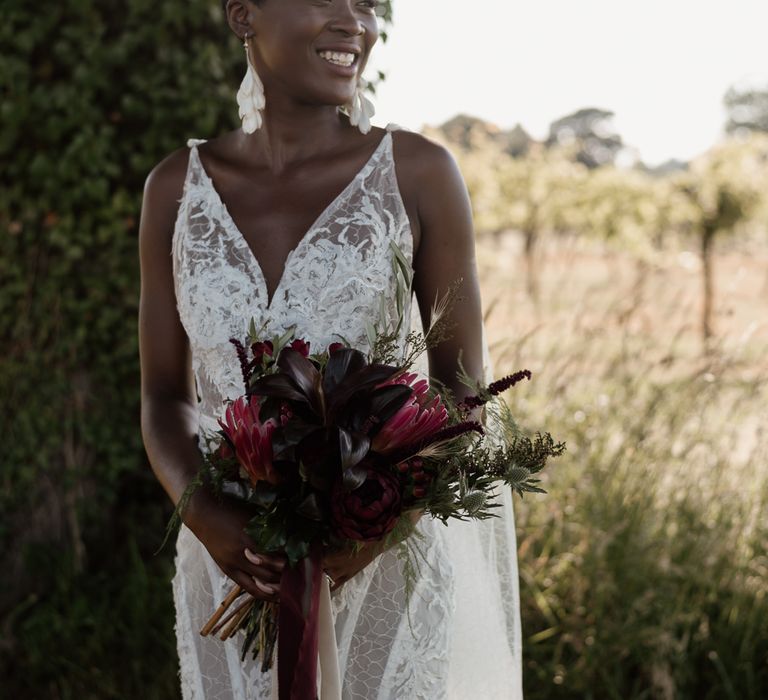 Black bride with cropped hair wearing a lace wedding dress and holding a king Protea wedding bouquet 