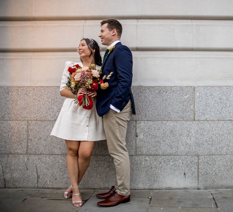 Groom in navy blazer embracing his bride in a short registry wedding dress holding a red and cream bouquet 