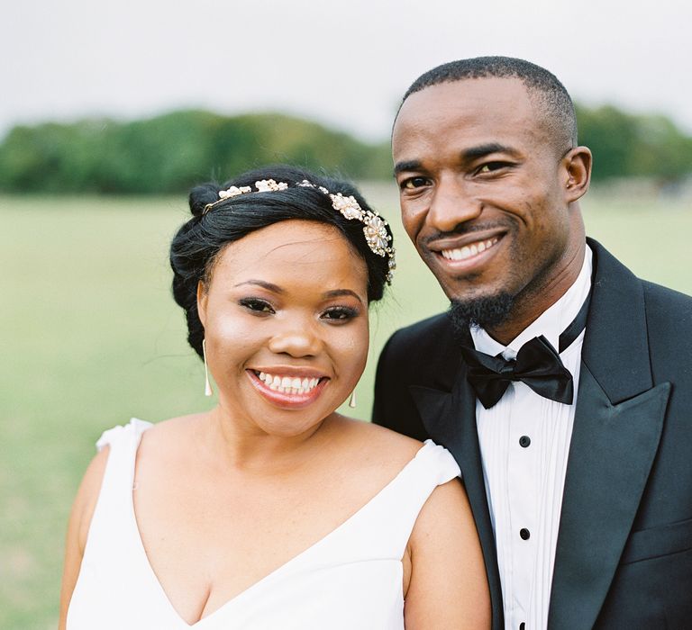 A Black bride and groom smile to camera. The bride wears her hair in an updo with a central parting and hairpiece. The groom wears a black tux.