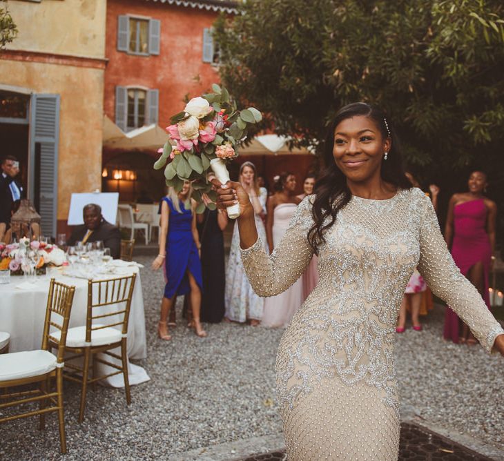 A Black bride in a long gold gown walks through her wedding with long soft waves in her hair and bouquet held high in her right hand 