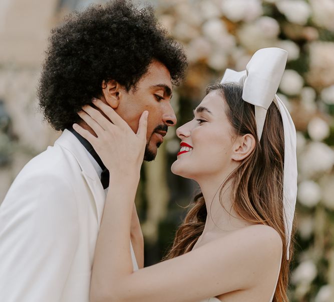 Bride with long brown hair, red lipstick and bow headdress kissing her black groom with afro hair and white tuxedo jacket 