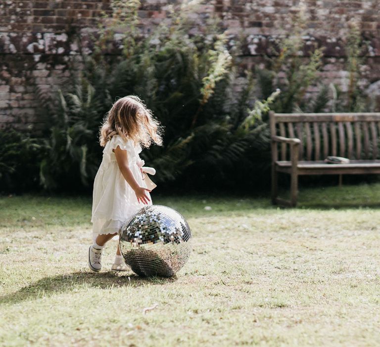 Flower girl in a white dress and trainers playing with a silver disco ball 