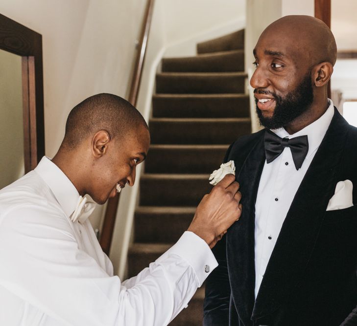 Groomsman pinning the Grooms white rose buttonhole onto his velvet tuxedo jacket 