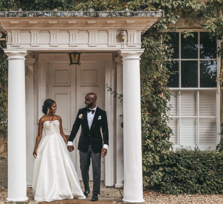 Black bride in a strapless Oleg Cassini wedding dress with embellished bodice holding her grooms hand in a velvet tuxedo outside Northbrook Park 