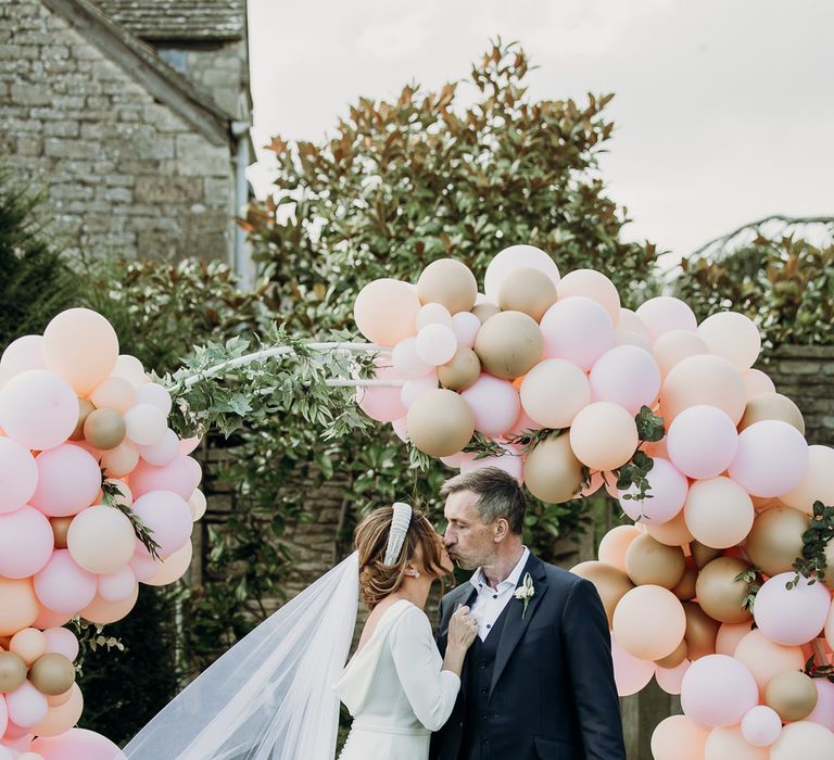 Bride in Elbeth Gillis gown kisses groom in navy Cad & the Dandy suit in front of pastel pink and gold balloon arch at Euridge Manor wedding