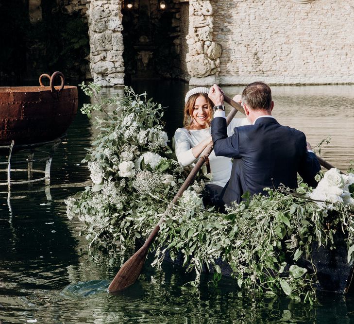 Bride in Elbeth Gillis gown sits with groom in row boat with floral installation on water at Euridge Manor Orangery wedding