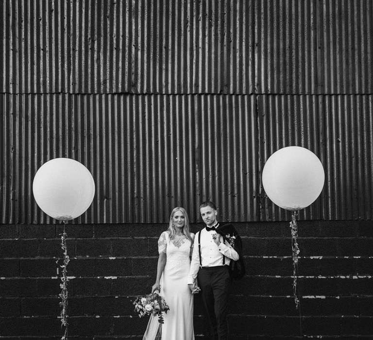 Bride in white Charlie Brear wedding dress holding rose and foliage wedding bouquet holding hands with groom in braces and bow tie at Caswell House wedding