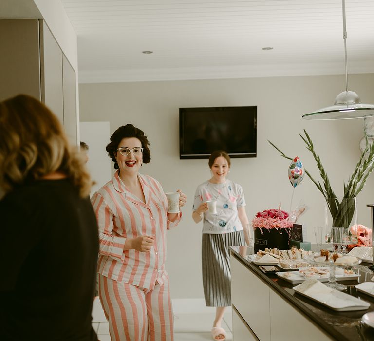 Bride in rollers and pink and white striped pyjamas holds a mug in a kitchen before the wedding