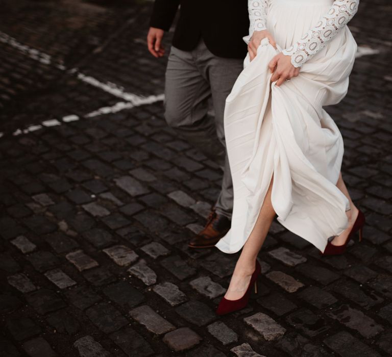 Bride in red suede heels and long sleeved lace top Self Portrait dress with pleated skirt walks with groom in grey trousers and brown shoes along cobbled street in Bristol