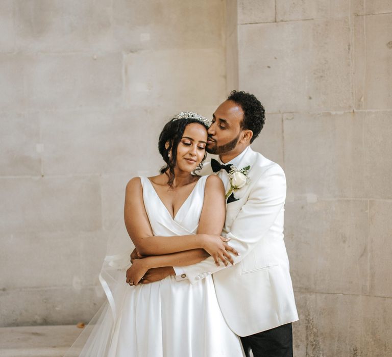 Groom in a white tuxedo jacket kissing his brides head in a princess dress and tiara 