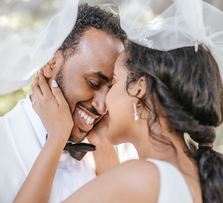 Bride with white nail polish caressing her grooms face under a wedding veil 