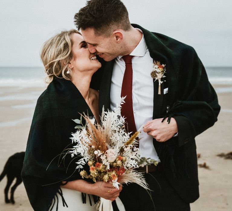 Bride & groom kiss whilst covered in shawl on the beach, as bride holds floral bouquet