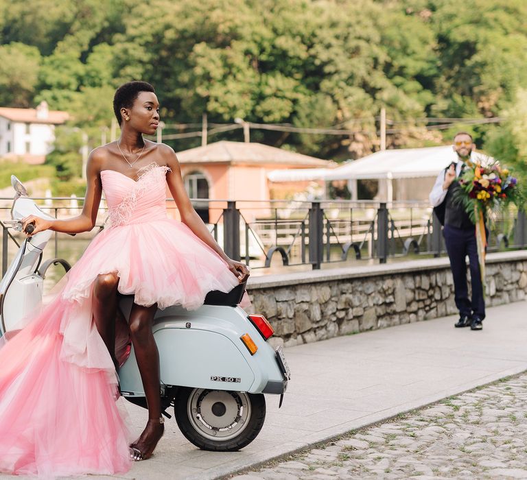 Bride sits on pale blue vespa beside Lake Como whilst wearing pastel pink bridal gown