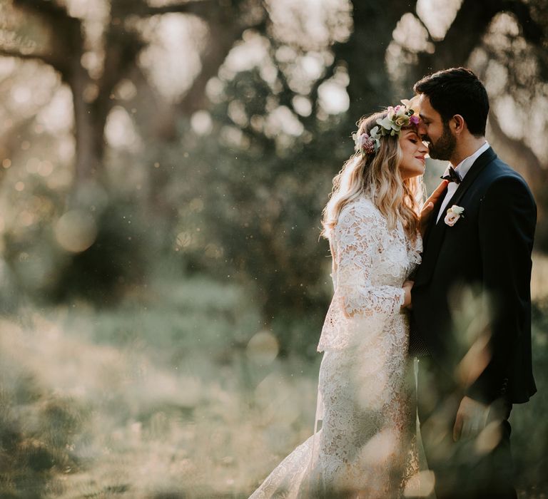 Bride in long wide sleeved lace wedding dress with curled hair and flower crown stands in forest with hands resting on groom in navy suit with bow tie at enchanted forest wedding