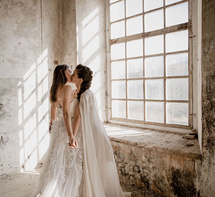 Bride in a ruffle skirt issing her bride with a braided updo and wedding cape by the window of an industrial venue 