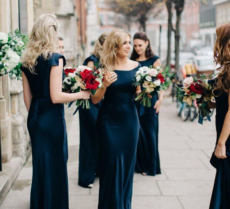 Bridesmaids in floor-length slip style navy wedding dresses with capped sleeves, holding red and white bouquets of roses | Ghost Dresses | M&J Photography