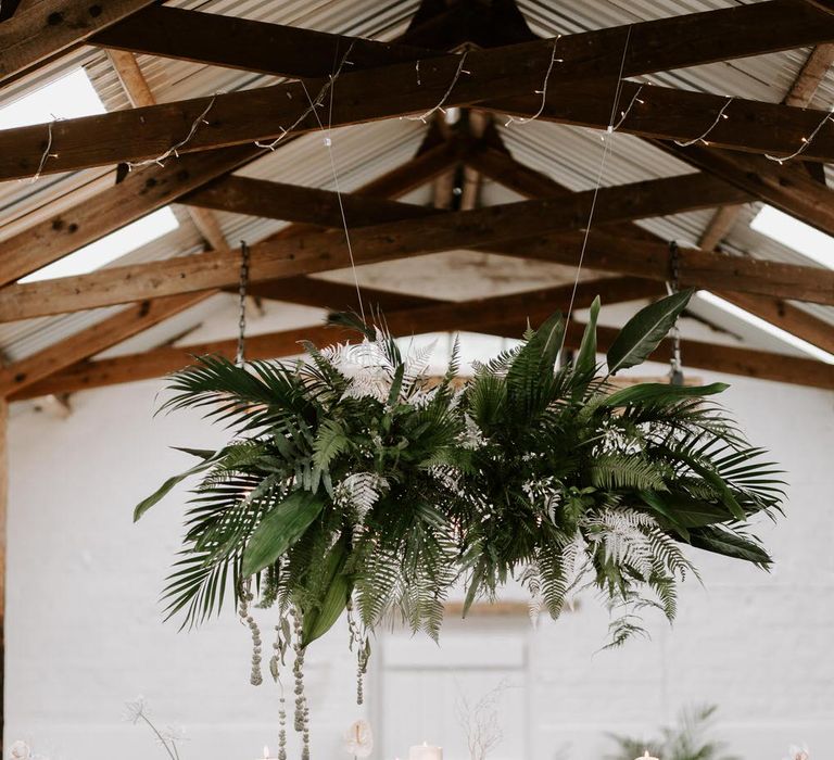 Tropical wedding table with industrial chic vibes and wooden bar stools, underneath a hanging installation of tropical leaves