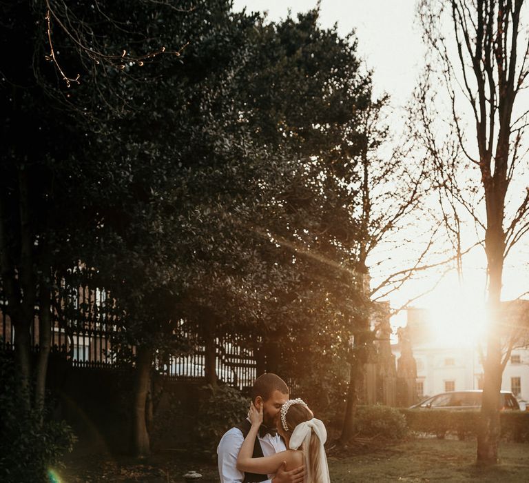 Golden hour portrait with the bride and groom kissing. The bride wears a long bow wedding veil 